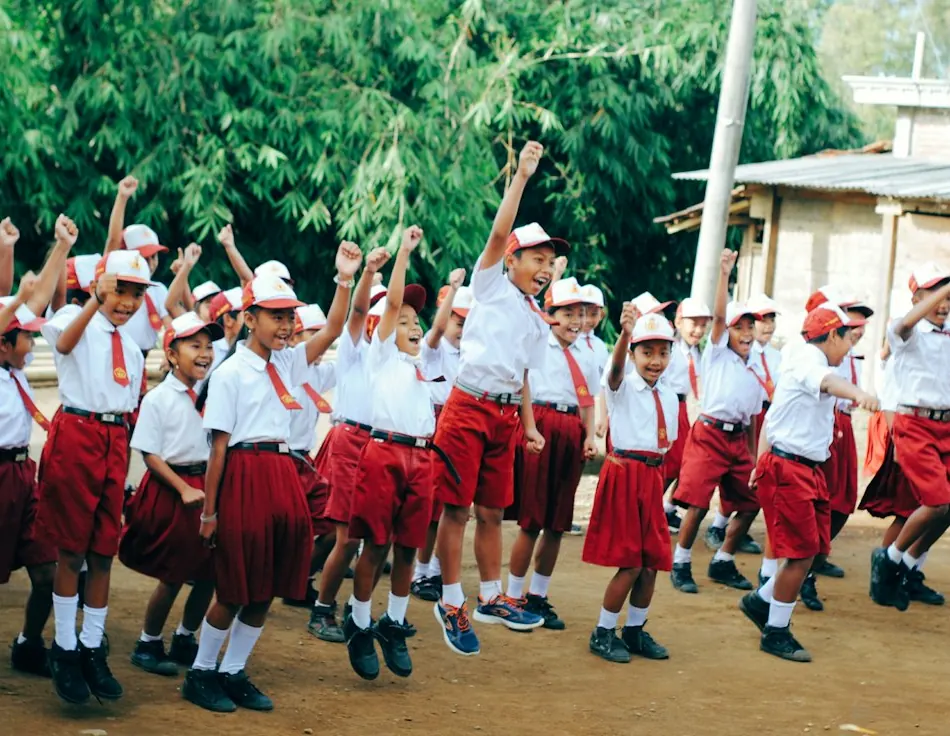 Children in white tops and red skirts or shorts, wearing white caps, raising their arms in a long line outdoors.