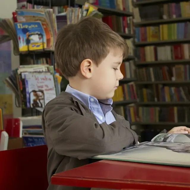 A boy reading an open book at a red table, with library shelves behind him.