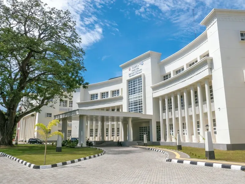 A white school building with tall columns and a curved paved forecourt, with a large tree on the left.