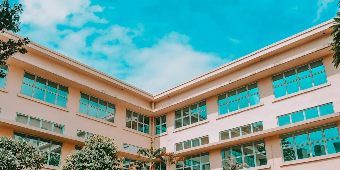A pink school building with teal-framed windows, seen from below against a blue sky with clouds.
