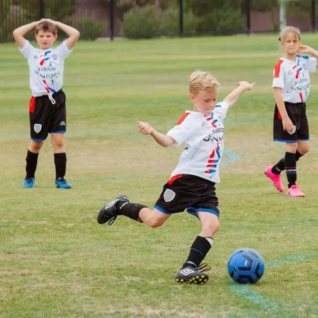 A boy in a white shirt kicking a football on a grass pitch, with two other children behind him.