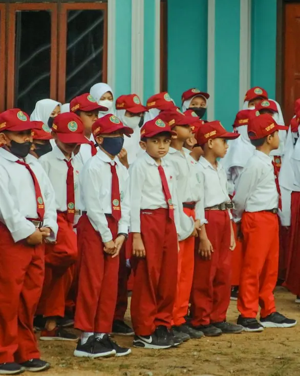 Rows of children in white shirts, red trousers and red caps standing in lines outdoors.