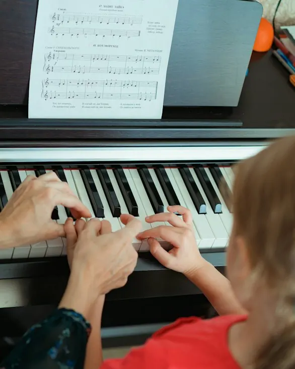 A teacher’s hands and a child’s hands on a piano keyboard, with sheet music above.