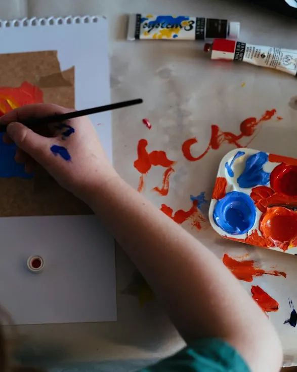 A child’s hand painting on paper beside a palette of red and blue paints.