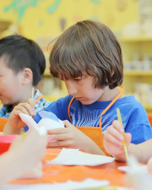 Children in aprons painting small clay pieces at a bright table.
