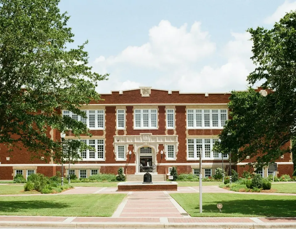 A red-brick school building with tall windows seen across a lawn, framed by two large trees.
