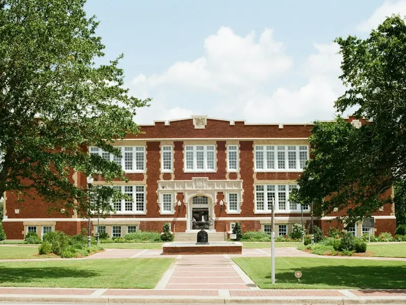 A red-brick school building with tall windows seen across a lawn, framed by two large trees.